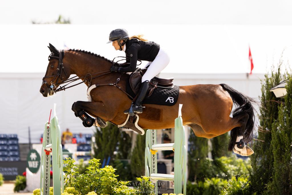 Ashlee and a bay horse clearing pale green rails